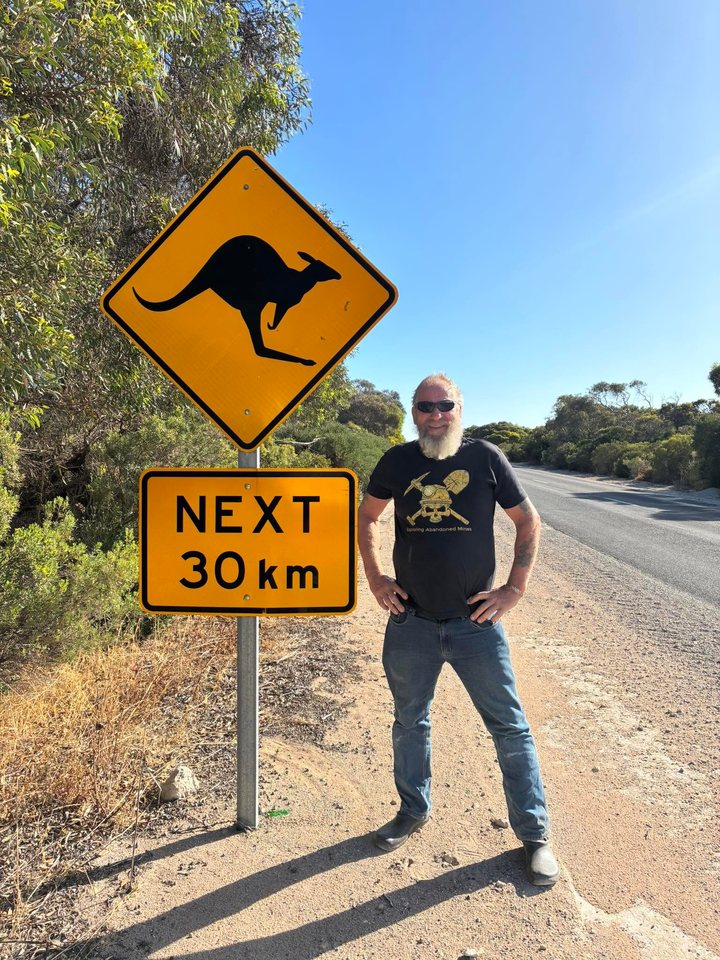 Frank Schlichting in sunglasses and an Exploring Abandoned Mines T-shirt, hands on his hips, beside a yellow kangaroo crossing sign at the edge of a road through the bush