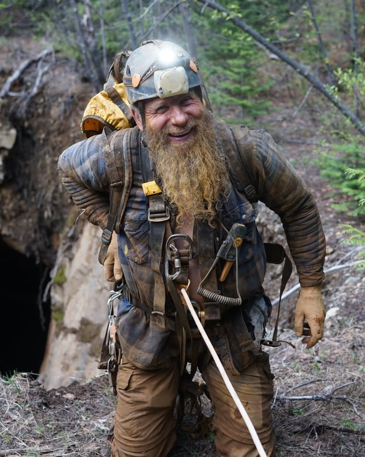 Frank Schlichting grinning through a long muddy beard as he climbs out of a mine opening on a rope, his headlamp still on
