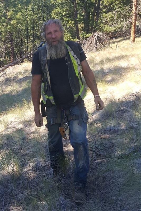 Frank Schlichting in a hi-vis vest and climbing harness, gear clipped at his waist, walking down a grassy slope among pine trees