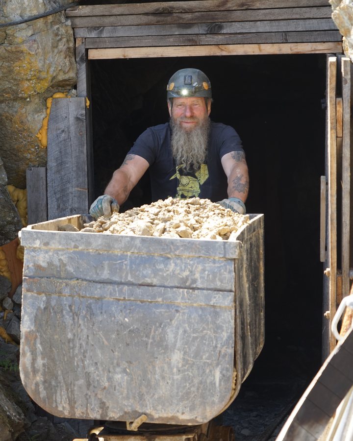 Frank Schlichting, smiling under his helmet and headlamp, pushing a steel ore car heaped with broken rock out of the timbered portal of the Yankee Boy