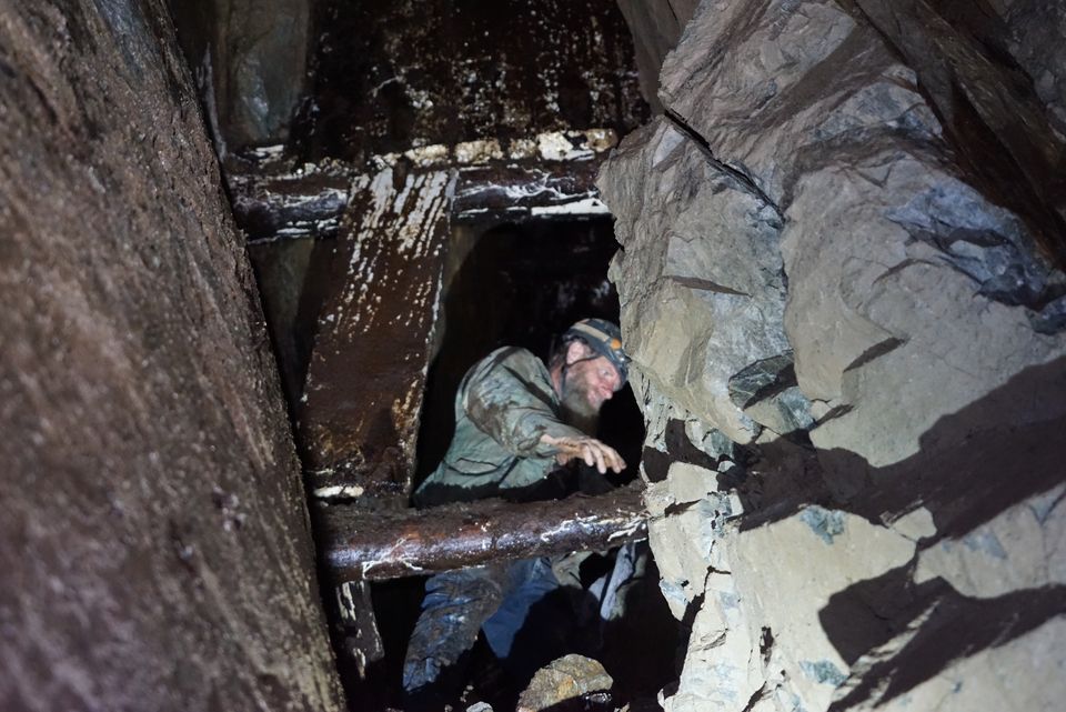 Frank in a muddy camouflage jacket and helmet, reaching through old wet timbers in a tight passage between walls of rock