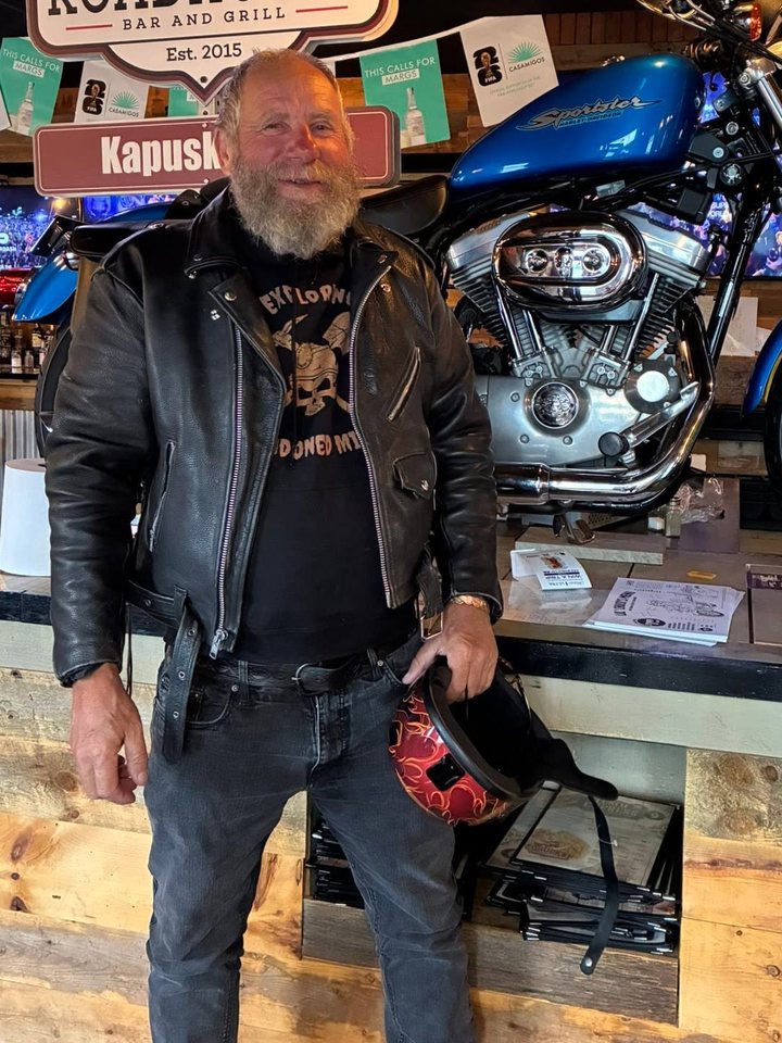 Frank Schlichting in a black leather jacket and an Exploring Abandoned Mines T-shirt, a flame-painted helmet in his hand, standing in front of a blue Harley-Davidson Sportster