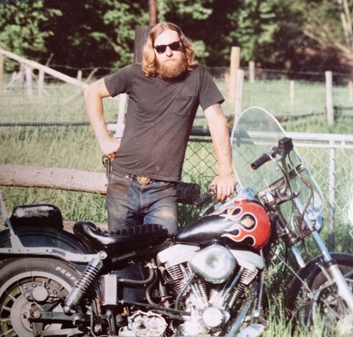 An old print of young Frank Schlichting, with long reddish hair, a full beard and sunglasses, leaning on a Harley-Davidson with flames painted on its tank, in a fenced pasture