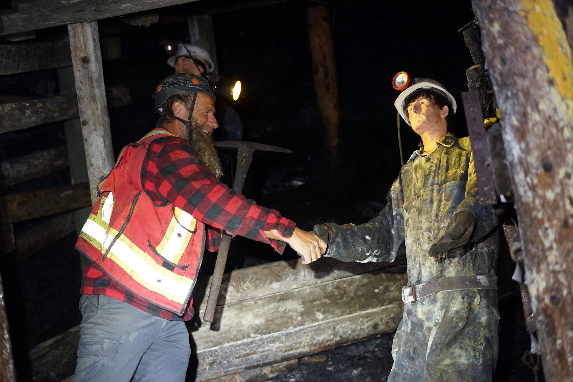 Frank Schlichting, grinning under his headlamp with a pick in one hand, shakes hands with a miner mannequin in an old timbered tunnel