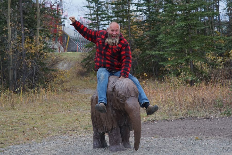 Frank Schlichting, laughing with one fist in the air, riding a statue of a baby woolly mammoth on a gravel path among spruce trees