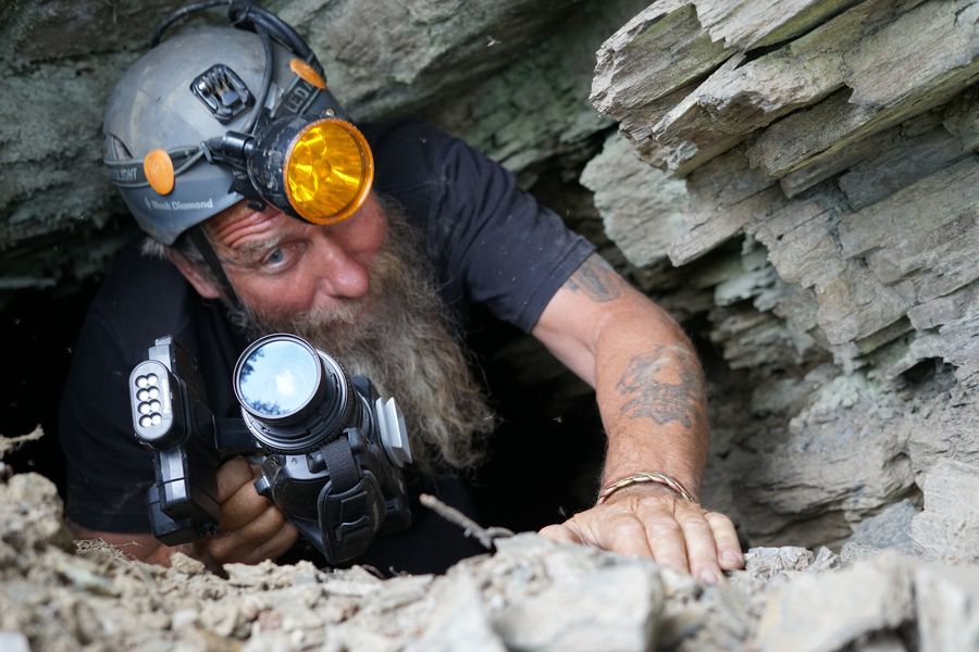 Frank Schlichting in a caving helmet with an amber lamp, filming with a camcorder as he squeezes through a narrow opening in layered rock