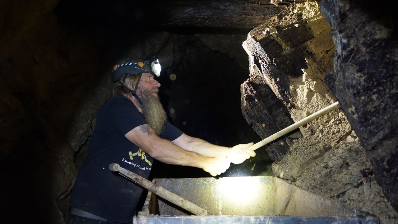 Frank Schlichting underground with his headlamp on, working a long wooden bar at an old timber chute above an ore car