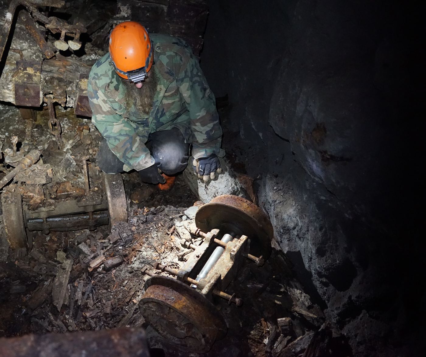 Frank Schlichting in an orange helmet and camouflage jacket, crouched by lamplight over old rusted mine machinery underground