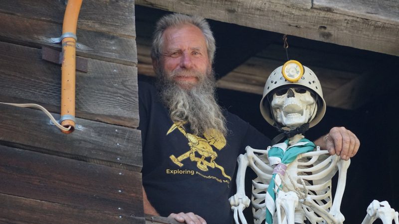 Frank Schlichting smiling from the window of an old wooden building, beside a skeleton in a miner&rsquo;s helmet and lamp