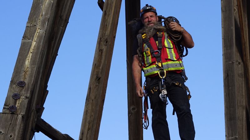 Frank Schlichting in a helmet, harness and hi-vis vest, a coil of rope over his shoulder, standing between the timbers of an old headframe against a blue sky