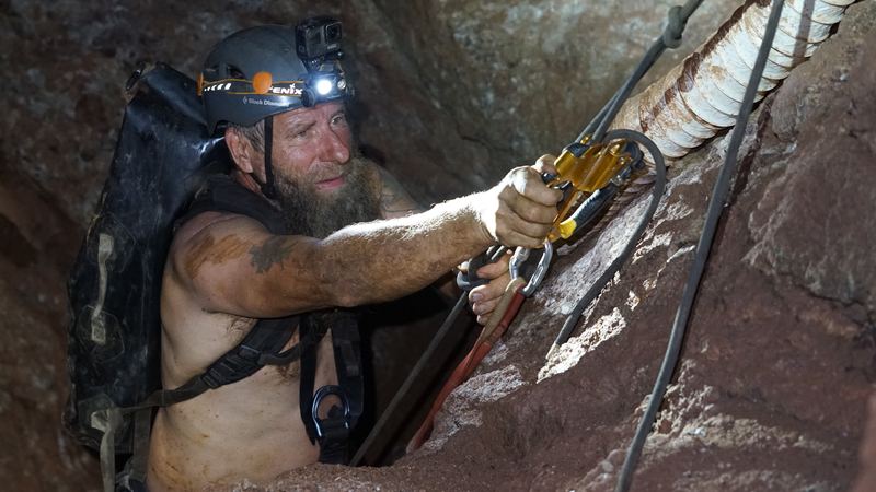 Frank Schlichting, shirtless in a helmet and harness, pulling himself up a rope beside an old pipe in a rock shaft