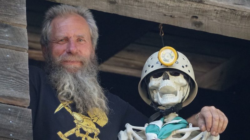 Frank Schlichting smiling from the window of an old wooden building, beside a skeleton in a miner&rsquo;s helmet and lamp