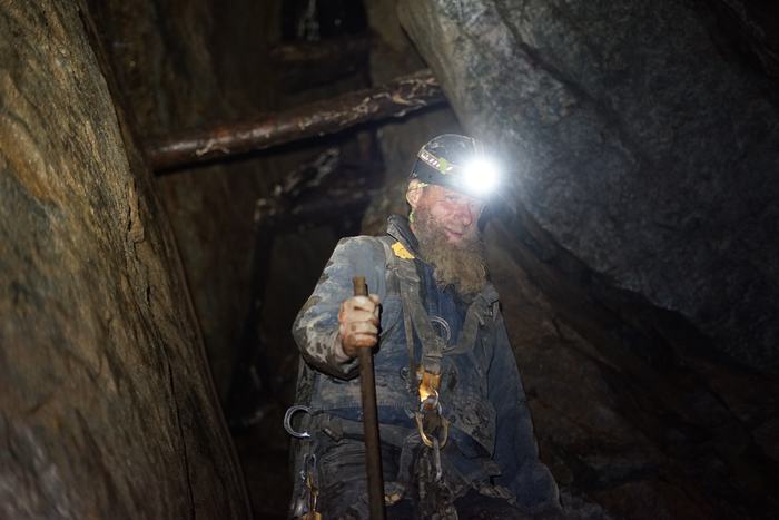Frank underground in a climbing harness, headlamp blazing, gripping a steel bar