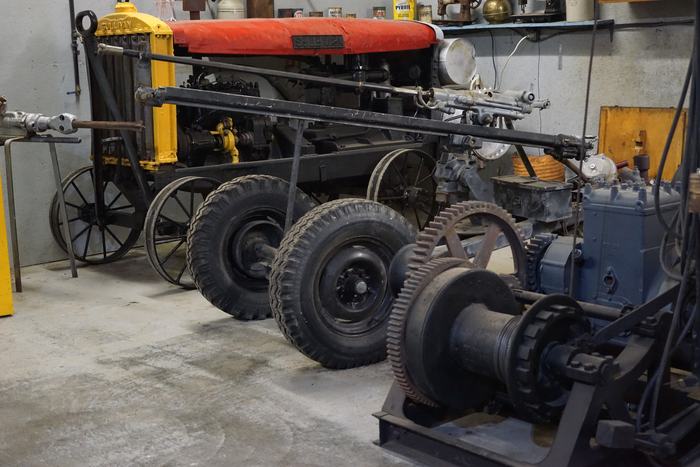 Old mining machines restored in a workshop: a Sullivan machine with a red hood and yellow radiator on iron wheels, rock drills on long bars, and a geared hoist