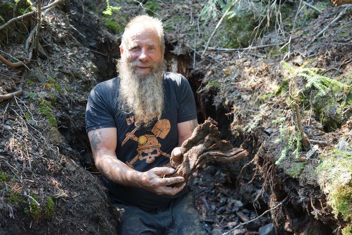 Frank smiling in a trench in the forest, holding up a rotted piece of old woven sacking
