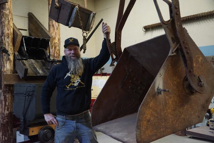 Frank in an Exploring Abandoned Mines hoodie, one hand up on the bail of a large rusted steel bucket in his workshop, with old ore cars and timbers behind him