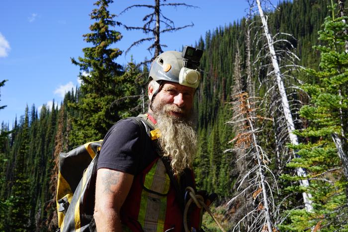 Frank outdoors in a helmet with a camera and lamp, a rope over his shoulder and a pack on his back, with a steep forested slope and blue sky behind him