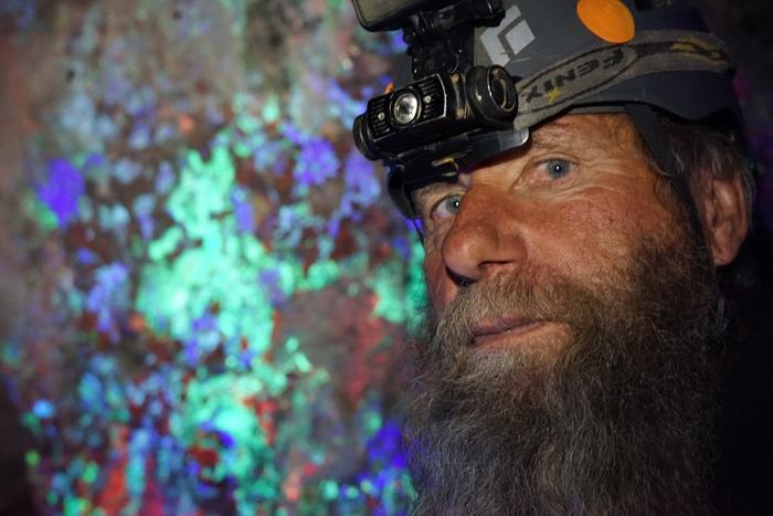 Close-up of Frank in his helmet, with minerals glowing green, blue and purple under ultraviolet light behind him