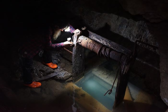 Frank leaning over an old wooden windlass wound with rusted cable, above a shaft full of clear blue-green water