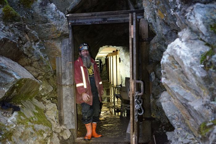 Frank Schlichting in a red work jacket and orange boots, smiling in the timbered doorway of the Yankee Boy mine, with lit timber sets running back into the tunnel