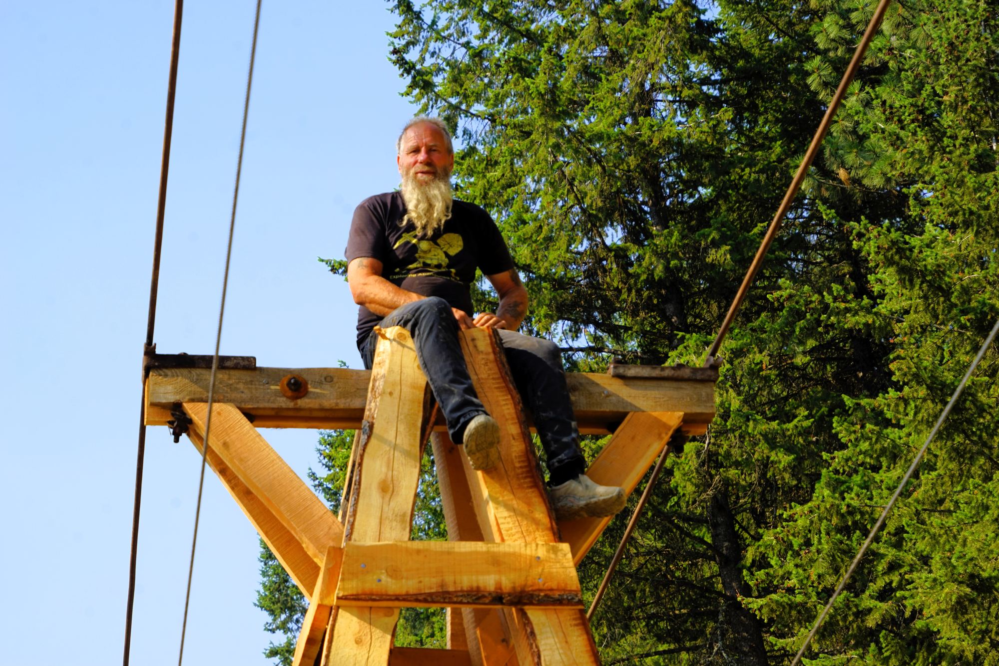 Frank Schlichting, grinning, sitting on top of a newly built timber tram tower, cables running past him, blue sky and tall spruce behind