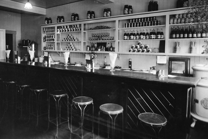 Inside the Bluebird Cafe at Anyox, BC: a long dark counter with a row of round stools, a coffee urn, pies and napkins on the counter, and shelves of tins, jars and bottles on the wall behind