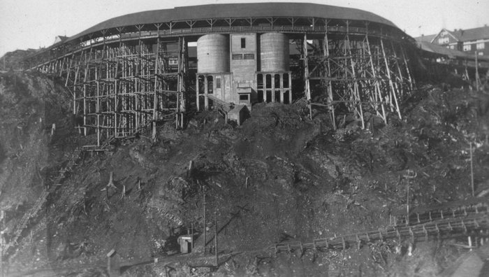 The concentrator at Anyox, BC: two concrete bins and a mill building on a rock slope, under a long curving timber trestle carrying the rail line, houses beyond