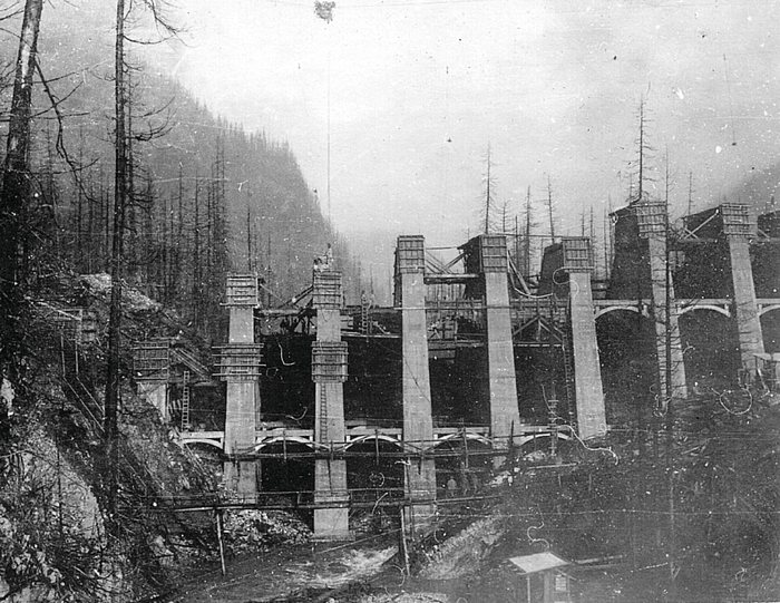 The concrete dam at Anyox, BC, under construction: tall concrete buttresses capped with wooden forms, arches rising between them, ladders and scaffolding, and bare, dead trees on the slopes above the creek