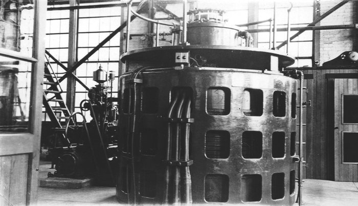 A large round generator in powerhouse No. 2 at Anyox, BC, a rail around it, a stair and tall windows behind