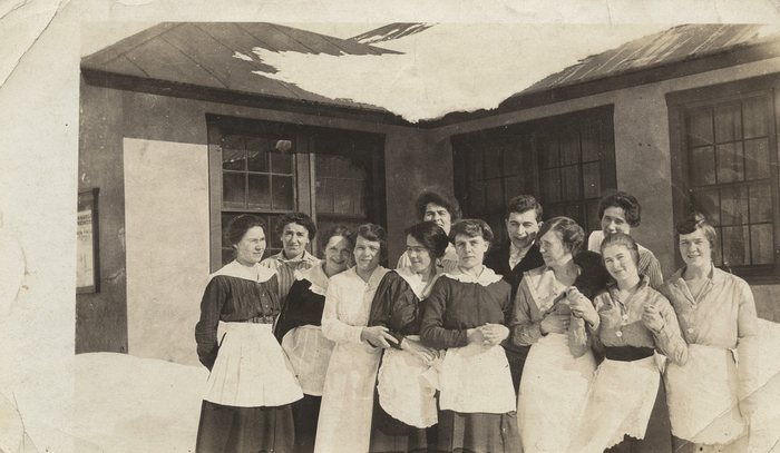 Hospital and nursing staff at Anyox, BC, most of them women in long white aprons, standing close together in the snow in front of a frame building, snow hanging from its roof