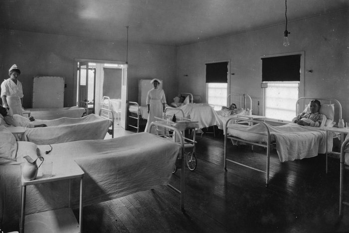 A hospital ward at Anyox, BC: two nurses in white uniforms and caps among iron beds, three men lying in them, a bare bulb hanging from the ceiling and the blinds half drawn