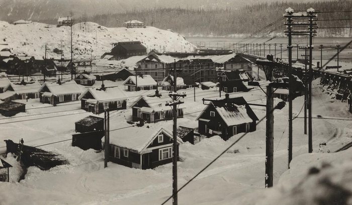 Company houses and larger frame buildings at Anyox, BC, buried in deep snow, power poles and wires in front and a trestle running along the water at the right