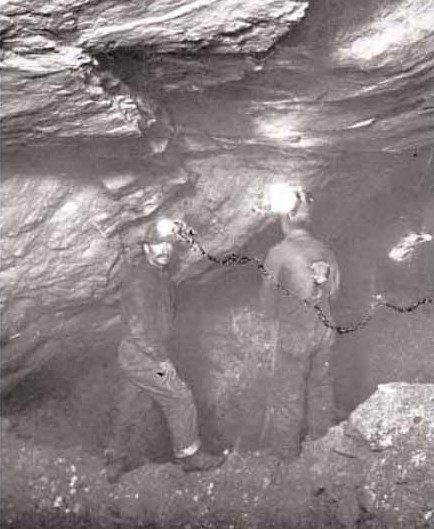 Two miners underground at the Hidden Creek mine, Anyox, BC, in 1917, lamps on their caps and a chain along the rock wall