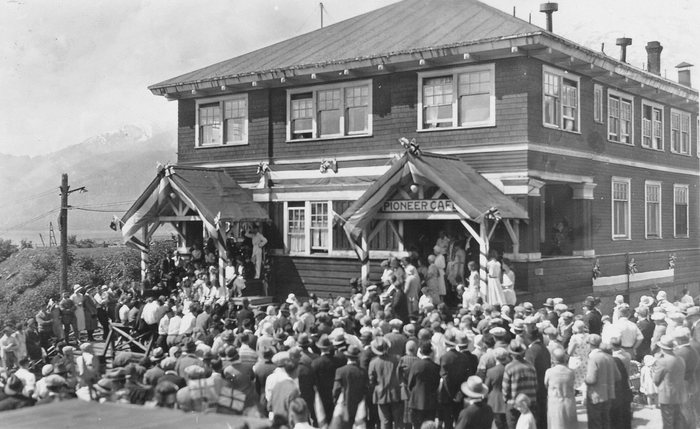 A large crowd in hats and coats gathered outside the two-storey Pioneer Cafe at Anyox, BC, its porches hung with Union Jacks and bunting, mountains beyond