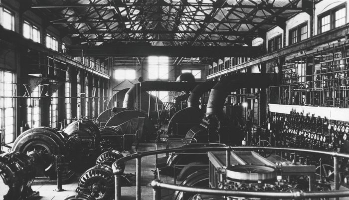 Inside powerhouse No. 2 at Anyox, BC: a long hall of turbines, generators and pipework under steel roof trusses and tall windows, a switchboard of dials along the right wall