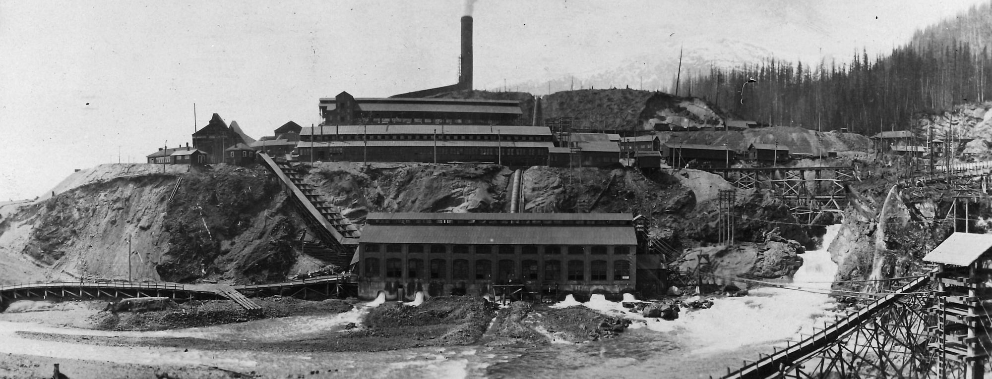 An old panorama of the works at Anyox, BC: the long powerhouse with arched windows at the foot of the hill, water pouring from beneath it into the creek and a waterfall at the right, and above it the smelter's long buildings and tall smoking stack