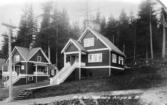 An old photograph of two new company houses at Anyox, BC, with porches and steep wooden stairs, a power pole and a plank walk in front and tall forest behind; printed on it, &ldquo;Residences Anyox B.C.&rdquo;