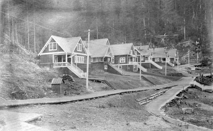 A row of company houses with porches and long wooden stairs on a hillside at Anyox, BC, plank walks crossing the cleared ground in front and tall forest behind