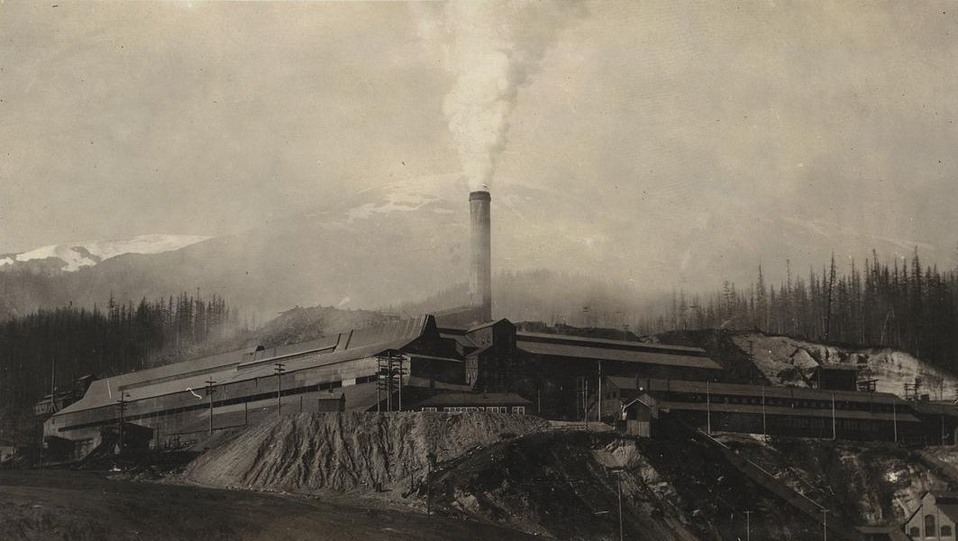 An old photograph of the smelter at Anyox, BC: long, dark smelter buildings along a slope under a tall stack pouring out a plume of smoke, a waste heap in front and a snow-capped mountain behind