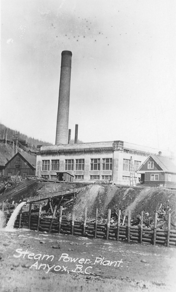 An old postcard of the steam power plant at Anyox, BC: a concrete building with tall windows under a very tall brick stack, a log crib wall and water spilling from a flume in front