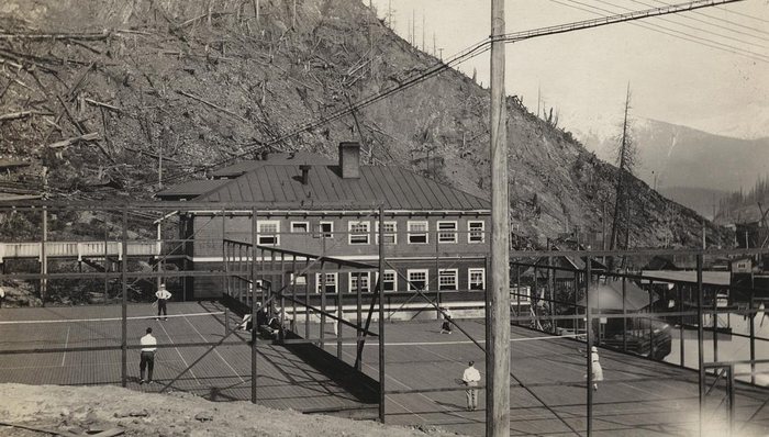 Players on wooden tennis courts at Anyox, BC, inside a frame of posts and wire, a large two-storey frame building behind them and a hillside of stumps and fallen trees above