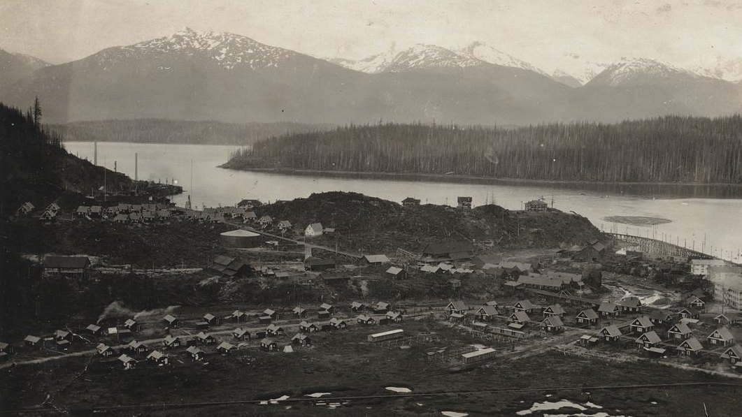 An old photograph looking down on Anyox, BC: rows of small frame houses on cleared, stump-strewn ground, a wharf trestle at the right, and across the inlet a forested point under snow-capped mountains