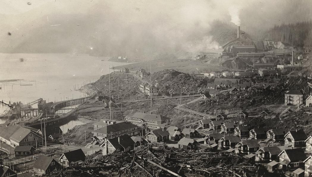 An old photograph looking over Anyox, BC: rows of houses on a slope strewn with stumps and fallen logs, the long wharf along the shore at the left, and the smelter beyond under a haze of smoke from its stack