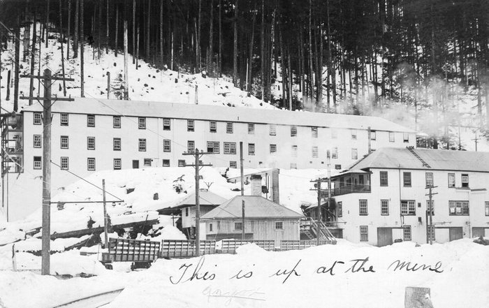 An old postcard of Anyox, BC, in winter: a long three-storey frame building below a forested slope, smaller buildings and a row of rail cars in deep snow, and a handwritten line, &ldquo;This is up at the mine&rdquo;