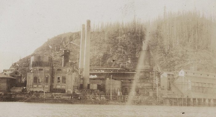An old, faded photograph of brick buildings with two tall stacks and a water tower at the water's edge at Anyox, BC, and behind them a hill of dead standing trees