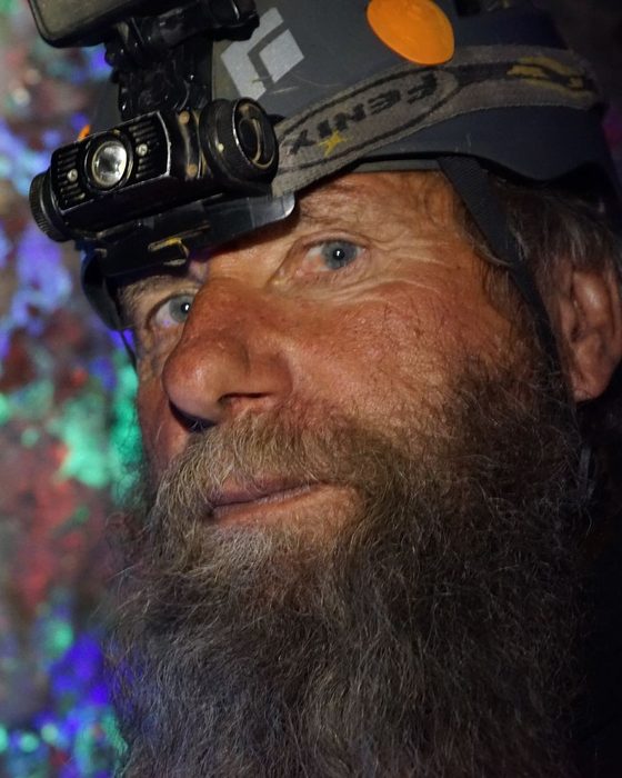 Frank Schlichting in his helmet and headlamp, close up, with minerals glowing green, blue and purple under ultraviolet light behind him