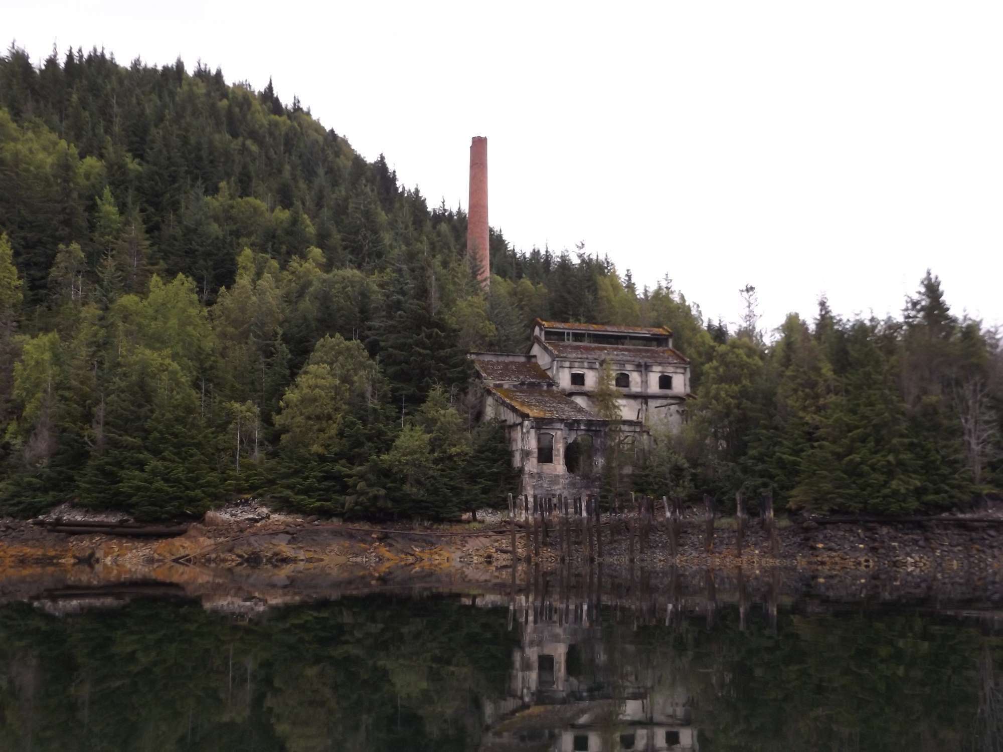 The ruined concrete powerhouse at Anyox, BC, a tall brick stack on the forested hill behind it and old pilings along the shore, all reflected in still water