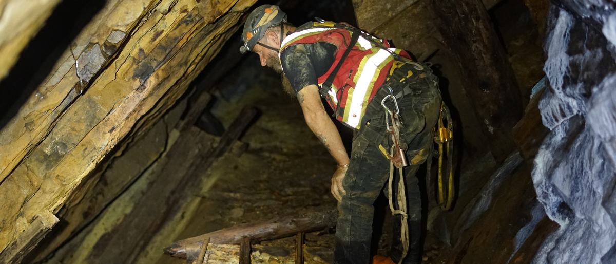 Frank Schlichting in a helmet, hi-vis vest and harness, stooping under old leaning timbers in a mine tunnel