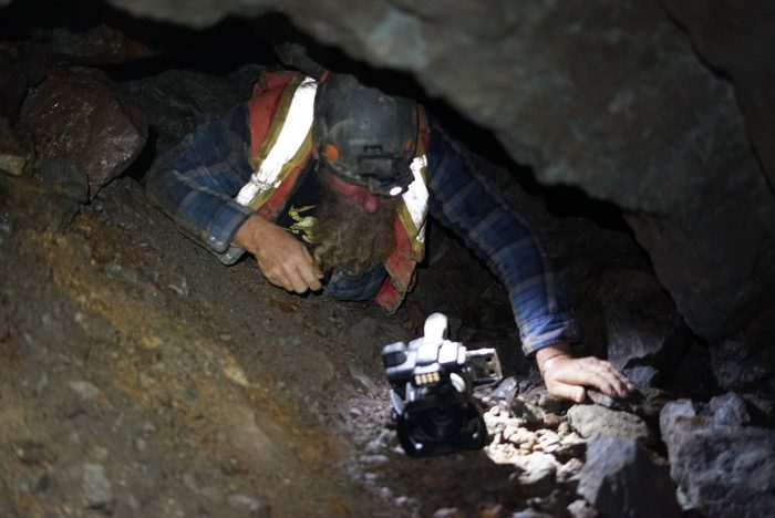 Frank crawling belly-down under a low rock back at Phoenix, pushing his camera ahead of him