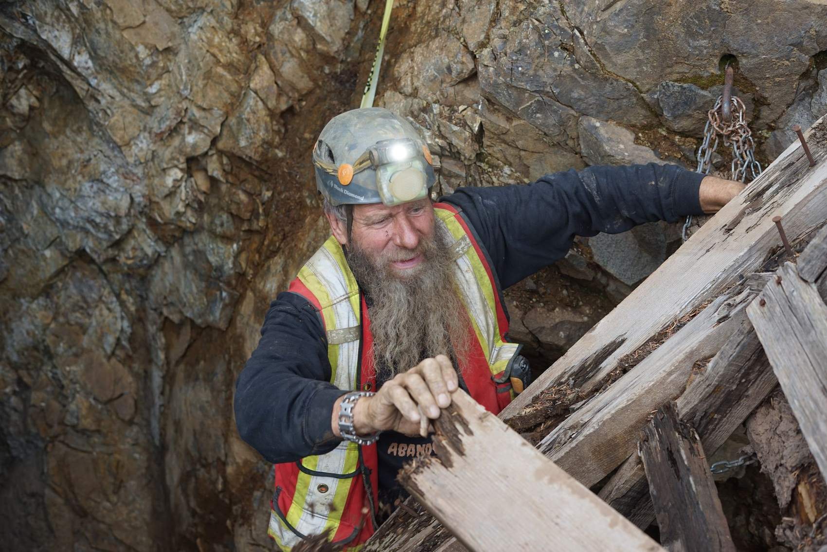 Frank Schlichting grinning in his helmet, headlamp and hi-vis vest as he climbs through broken stope timbers in the old workings at Phoenix, BC
